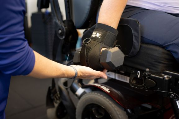 Image shows female clients hand with an Active Hands grip device holding a dumbell. Pauline is helping to guide the dumbell.