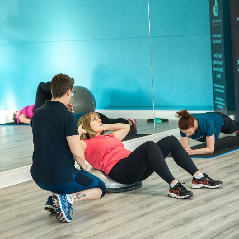 Pauline is crouched beside a female client who is performing an ab crunch. You can see other class members in the background.