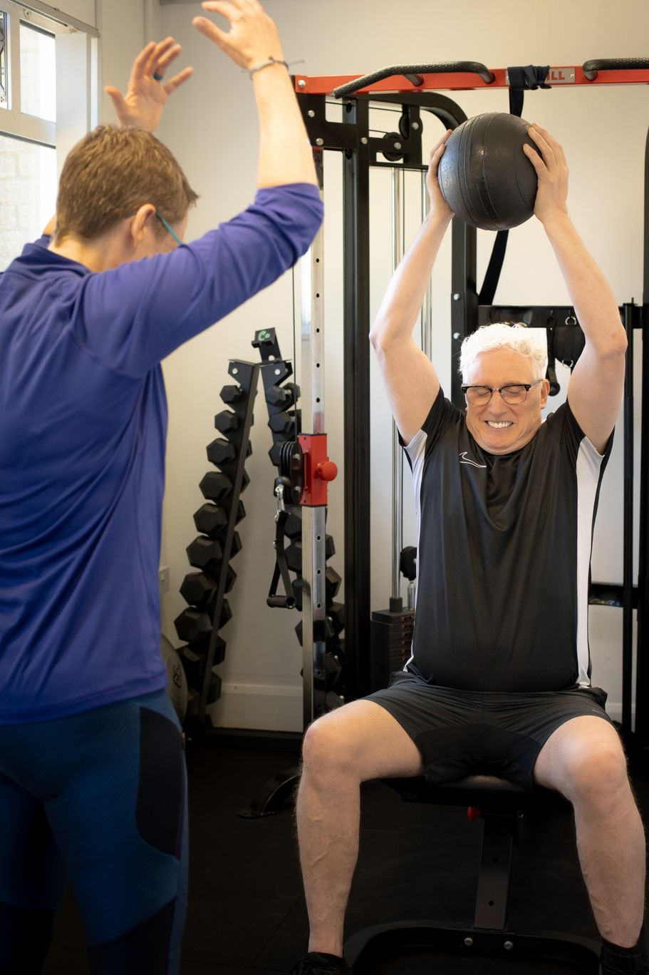 Trainer demonstrating an exercise with male client seated holding a slam ball above head