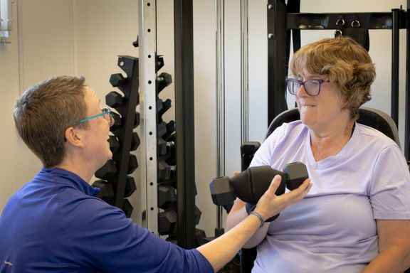 Client is performing a bicep curl being assisted by Pauline and using an active hands gripping device to hold the weight.