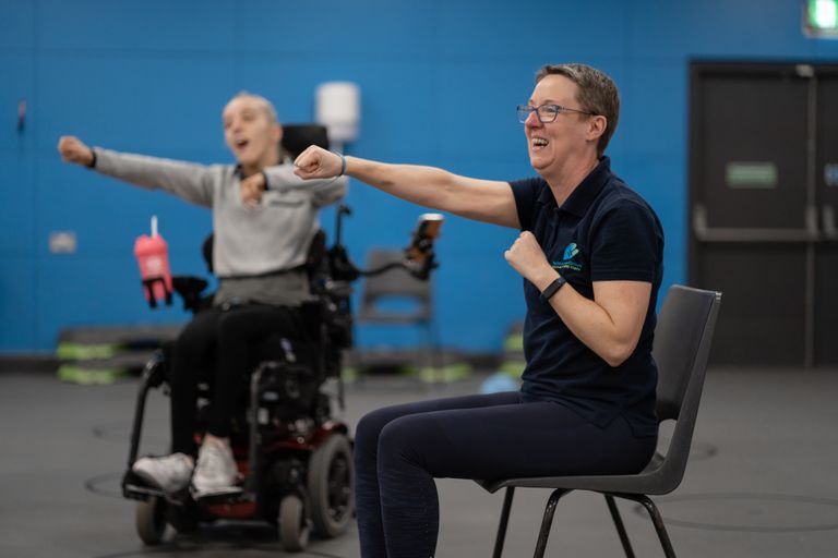 Paulne is seated and doing an right arm punch movement. A class participant in an electric wheelchair is copying the move. Both are smiling.