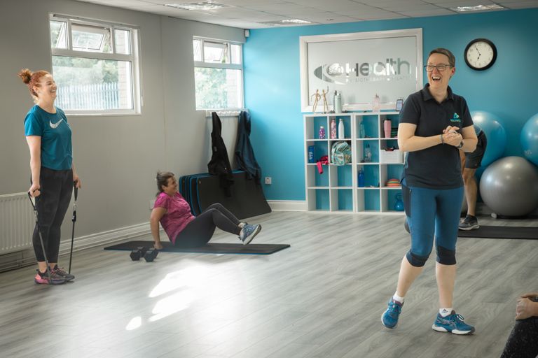Pauline is standing with two class participants exercising, one doing a bicep curl and the other on the floor. All are laughing.