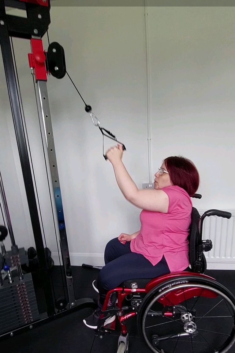 A female client in a manual wheelchair is reaching above her head with her left arm to pulldown a weight on a cable machine.